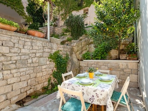 A stone-walled outdoor dining area with a table set for two, surrounded by greenery and a stone wall. - Beautiful Holiday Home on the Island Brac, With the Sea and a Small Beach in Front of the Door (Pucisca)