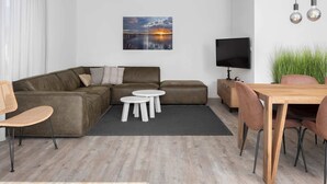 A modern living room with a sectional sofa, a small round table, a TV, and a dining area with chairs. - Villa by Lake Veere With Sauna (Wolphaartsdijk)