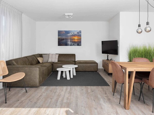 A modern living room with a sectional sofa, a small round table, a TV, and a dining area with chairs.