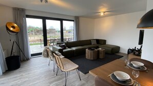A modern living room with a sectional sofa, a dining table set for two, and a large window offering a view of the outdoors. - Villa by Lake Veere With Sauna (Wolphaartsdijk)