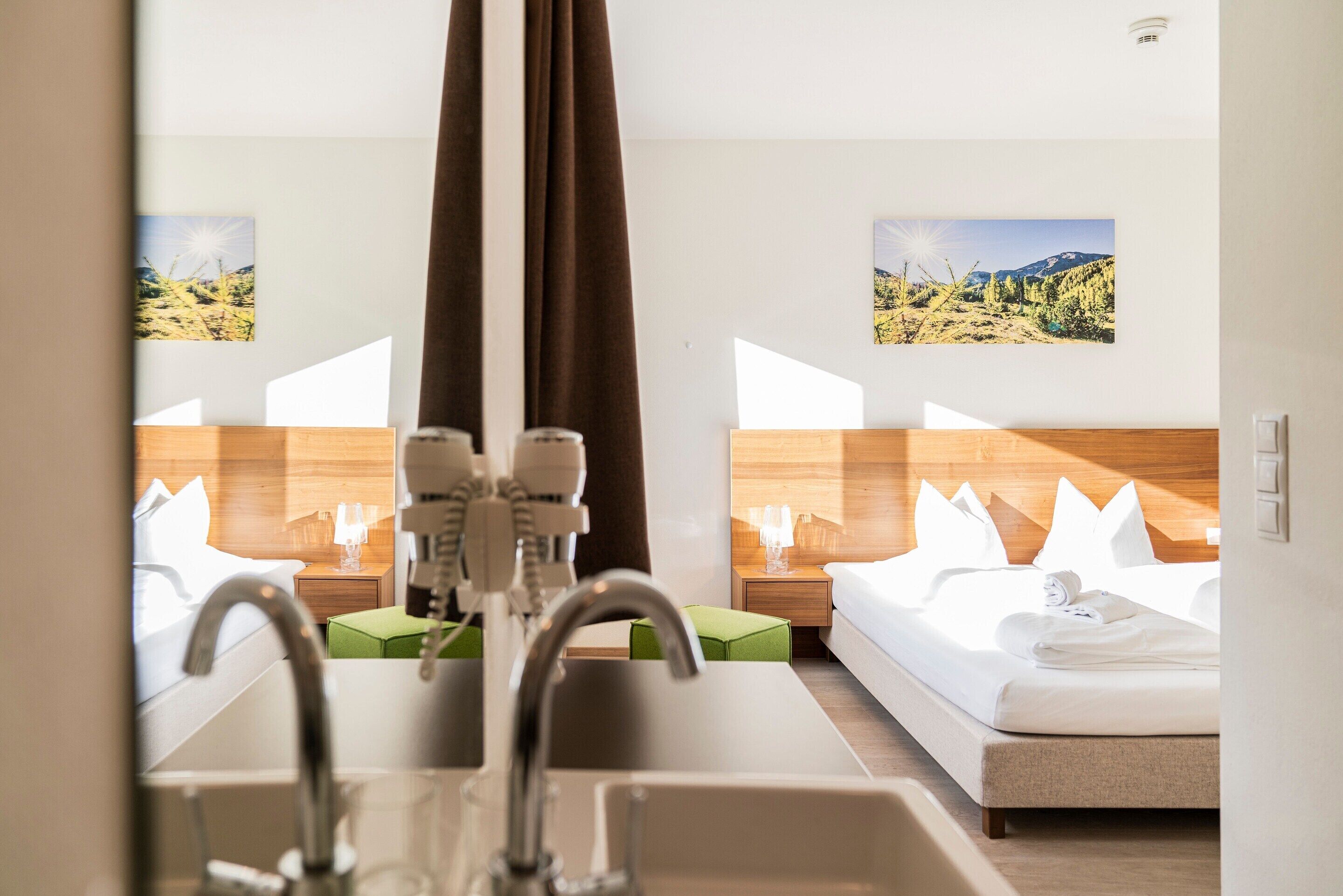 A hotel room with a modern bathroom, a bed with white linens, a nightstand with a lamp, and two landscape paintings on the wall.