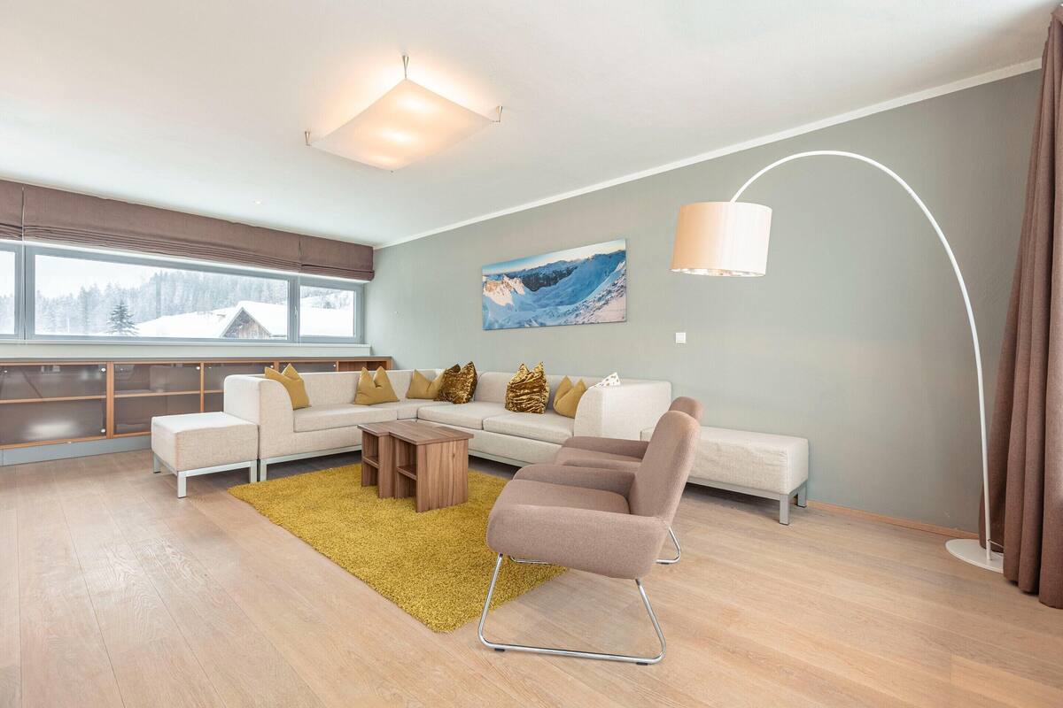 Un salon moderne avec un canapé d’angle, une table basse et un lampadaire. Une grande fenêtre offre une vue sur un paysage enneigé.