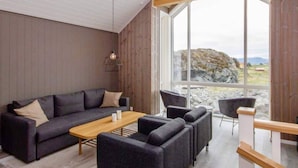 Een moderne woonkamer met een zithoek, een salontafel en een eettafel. Een groot raam biedt uitzicht op het omliggende landschap.