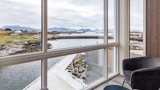 Een kamer met een groot raam met uitzicht op een fjord, een houten terras en een stoel.