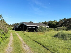 A single-story building with a sloped roof, surrounded by a grassy field and dense trees. - Holiday Home in LÃ¸kken (Lokken)