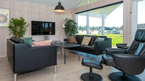 A modern living room with a sofa, armchair, and TV, wooden walls, and large windows. - Holiday Home in Nørre Nebel (Norre Nebel)