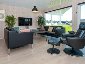 A modern living room with a sofa, armchair, and TV, wooden walls, and large windows.