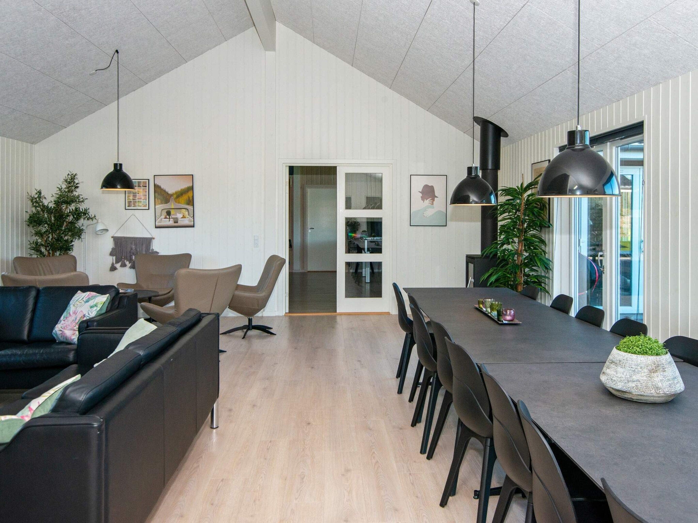 A modern living room with a dark sofa, a coffee table, and a dining area with a long table and chairs. There are pendant lights above the dining table and a large window with a view.