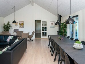 En moderne stue med en mørk sofa, et sofabord og et spiseområde med et langt bord og stole. Pendellamper hænger over spisebordet, og et stort vindue giver udsigt.