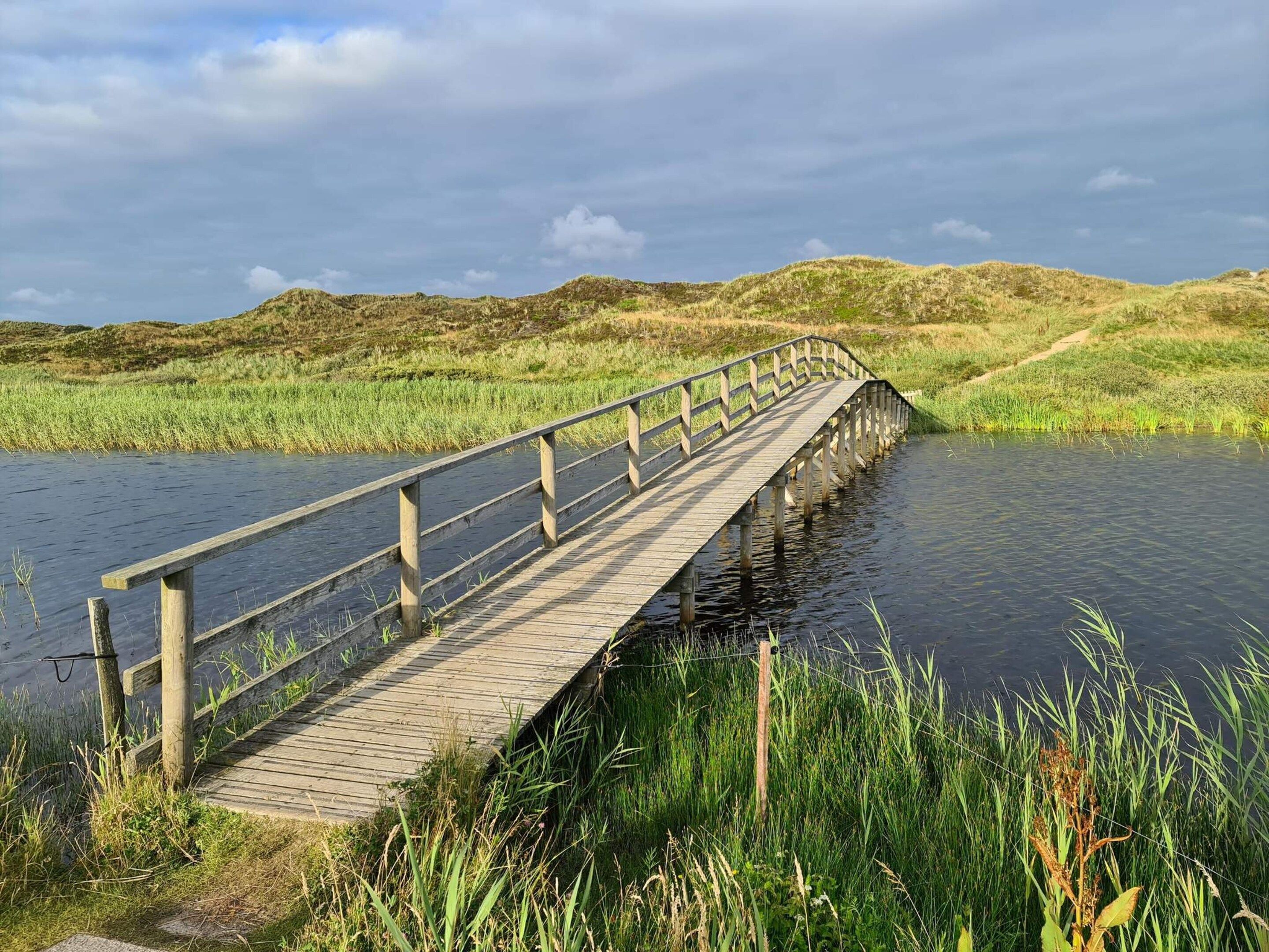 A wooden bridge over a body of water with grassy banks and hills in the background.