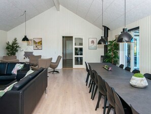 A modern living room with a dark sofa, a dining area with a long table and chairs, and a large window with a view. - Holiday Home in Nørre Nebel (Norre Nebel)
