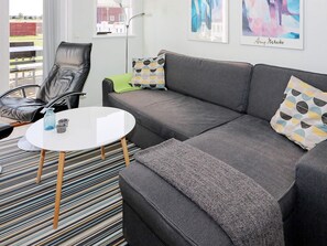 Un salon moderne avec un canapé gris, un fauteuil en cuir noir, une table basse blanche et un tapis à rayures. Deux œuvres d’art sont accrochées au mur, et une fenêtre offre une vue sur un bâtiment rouge.