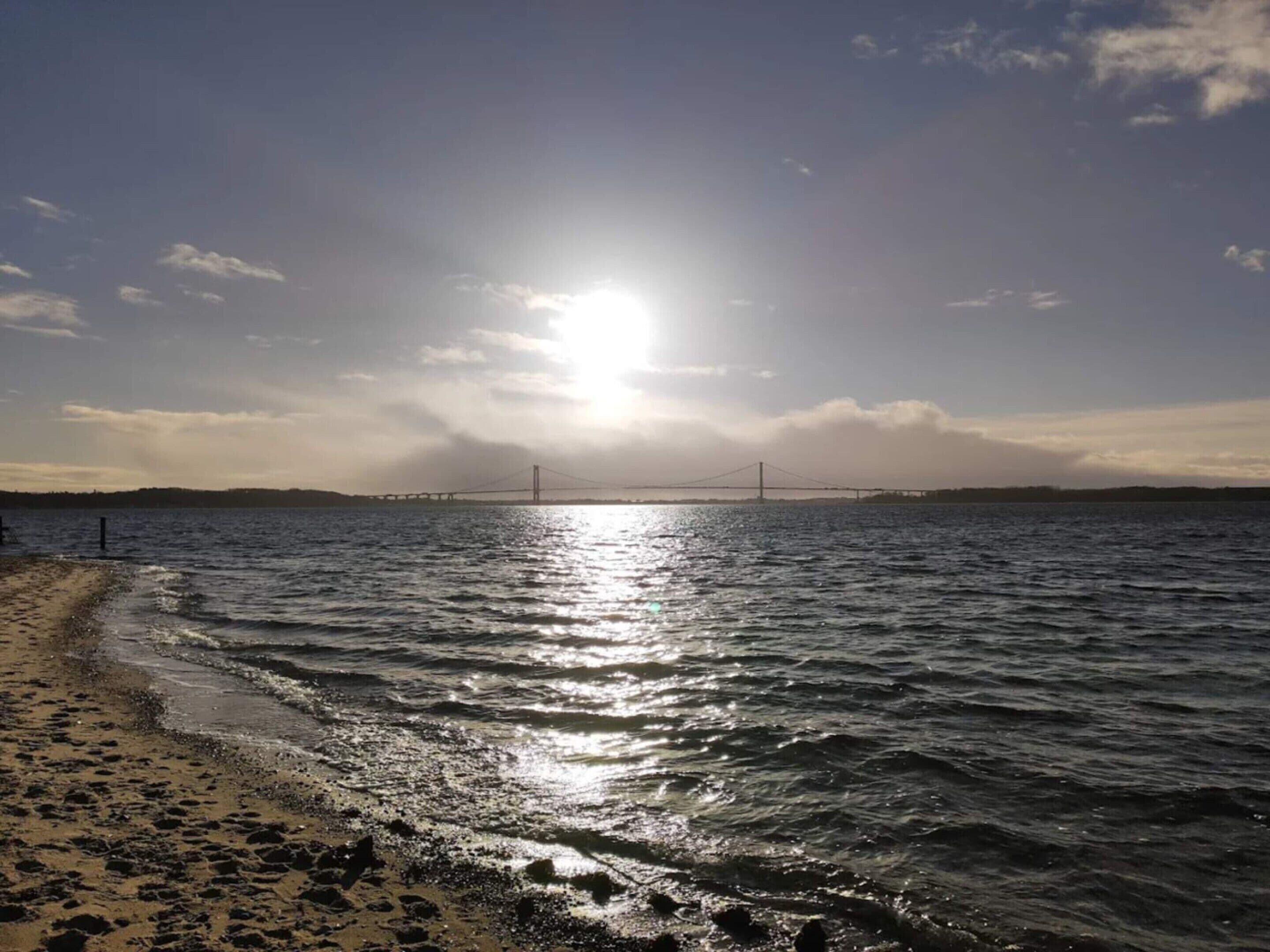 A beach with a bridge in the distance, calm water, and a clear sky.