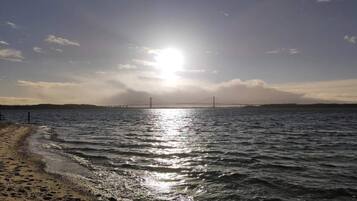A beach with a bridge in the distance, calm water, and a clear sky.
