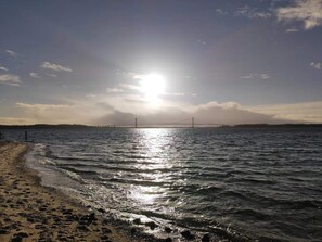 A beach with a bridge in the distance, calm water, and a clear sky.