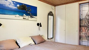 Een slaapkamer met een bed, een schilderij van een strandgezicht, een spiegel en een deur met een decoratief glasraam.