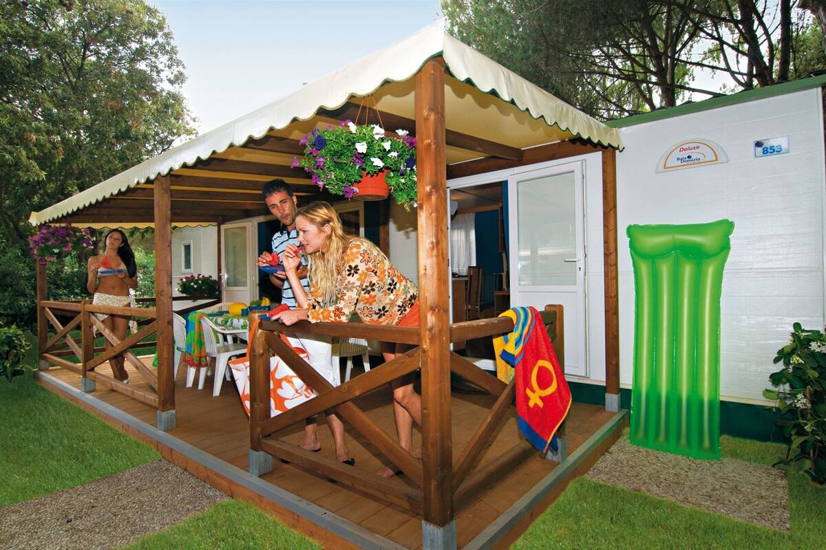 Una terraza de madera con un área cubierta, una piscina inflable verde y un edificio blanco con una puerta.