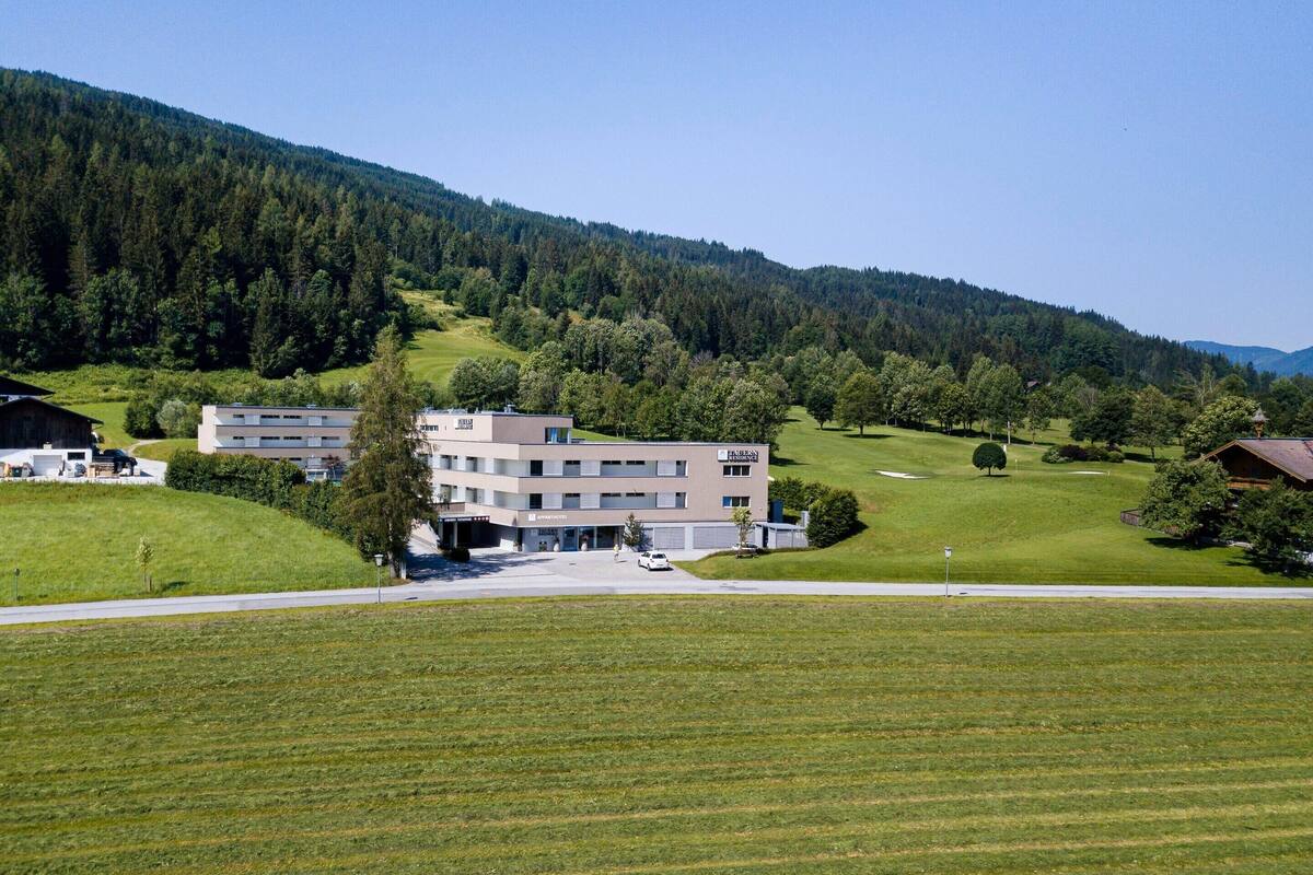 A modern building with multiple floors, surrounded by a green landscape and a forested hill in the background.