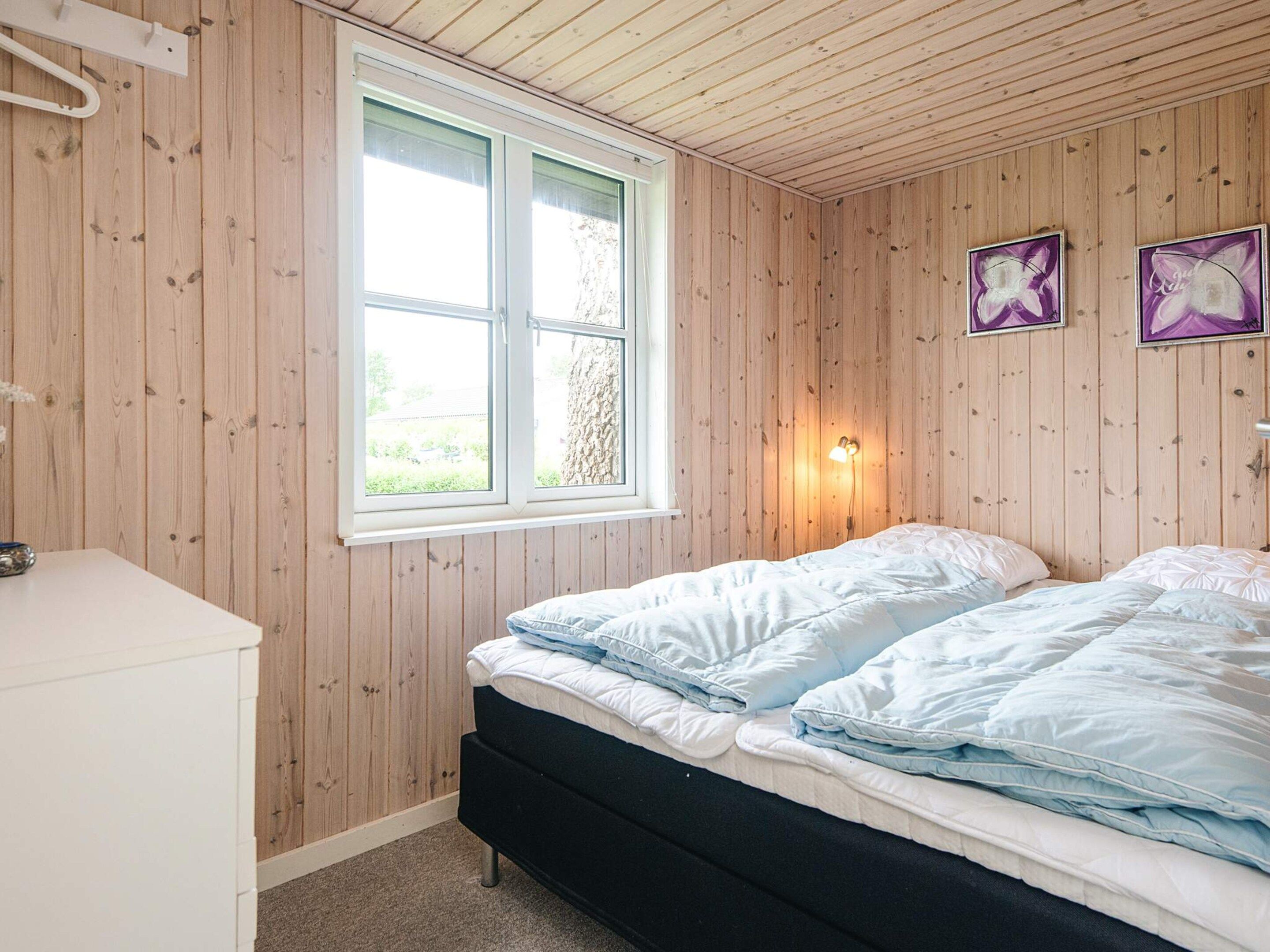 Ein Schlafzimmer mit einer holzverkleideten Wand, einem Doppelbett mit weißer und blauer Bettwäsche, einem weißen Nachttisch und einem Fenster mit Blick auf Grünflächen.