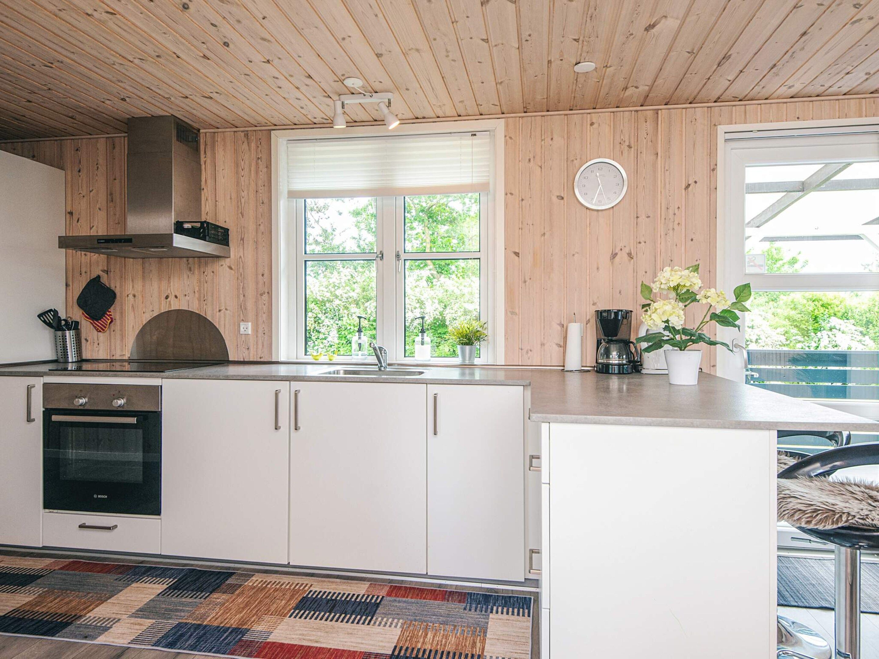 A modern kitchen with white cabinets, a stainless steel range hood, a built-in oven, a large window with a view of greenery, and a patterned rug.