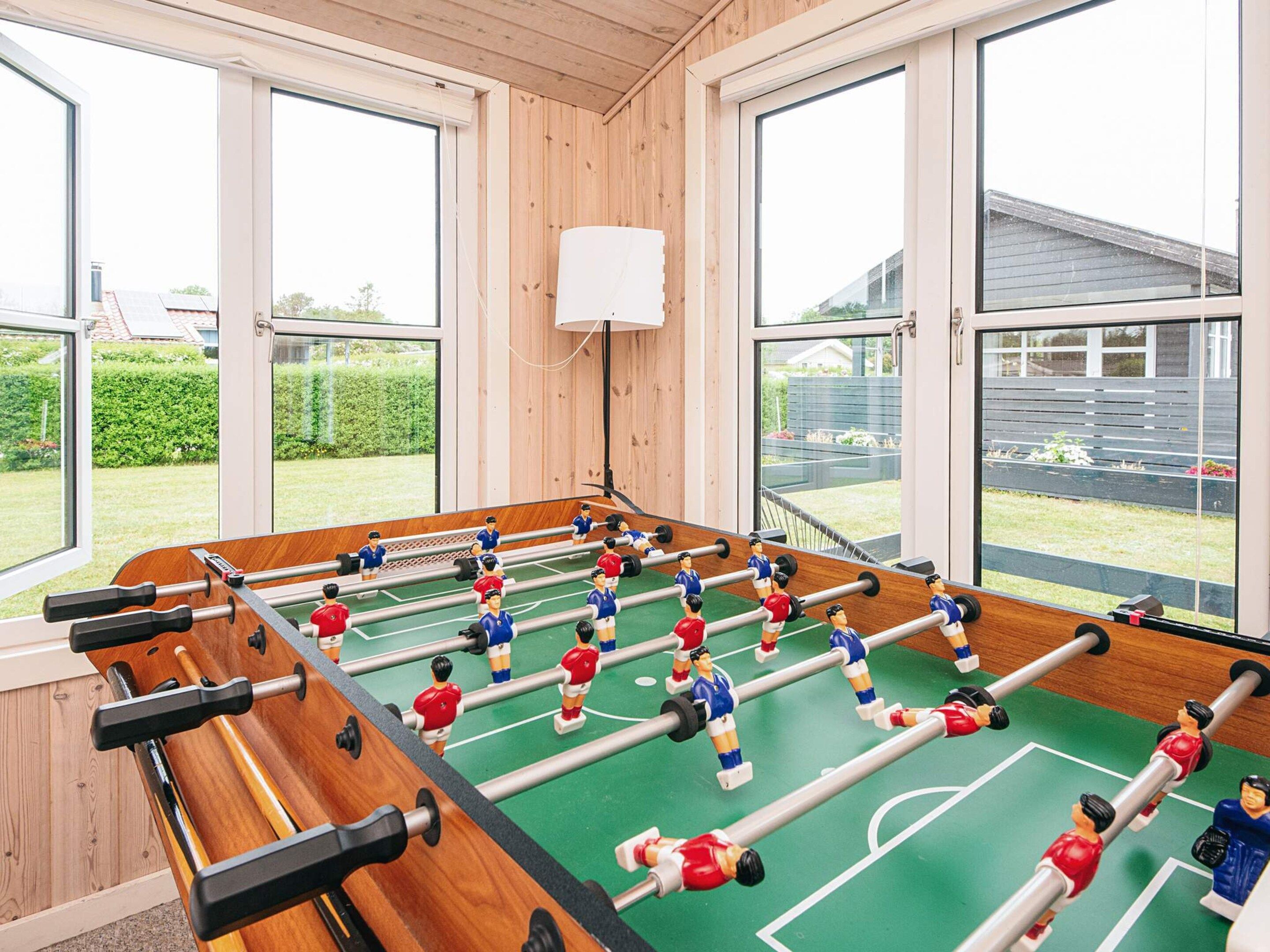Ein Kicker mit Holztisch und einer grünen Spielfläche, platziert in einem Raum mit großen Fenstern und einer weißen Decke.