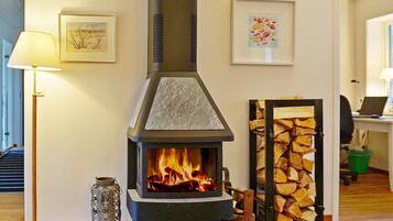 A room with a wood-burning stove, a stack of firewood, and two framed artworks on the wall.