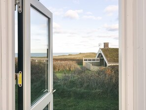 A view through a window of a coastal landscape with a grassy field, a small building with a sloped roof, and a body of water in the distance.