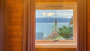 Una habitación con un amplio ventanal que da a un cuerpo de agua y montañas, paredes de madera y una zona de estar sencilla.