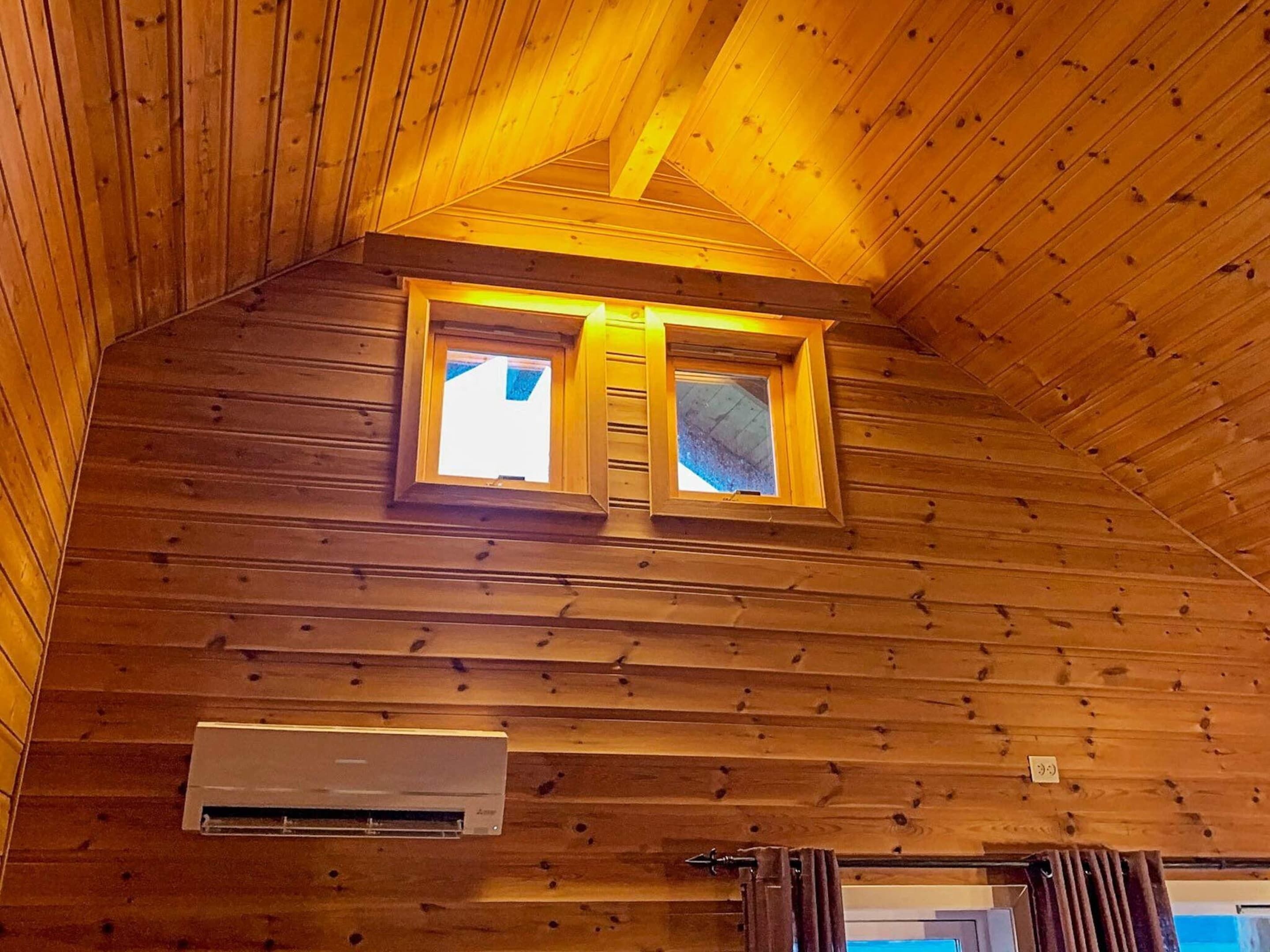 A wooden cabin interior with a sloped ceiling, two windows with a ladder, and an air conditioning unit.