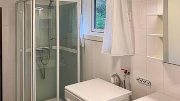 A modern bathroom with a glass-enclosed shower, a built-in washing machine, and a window with a view of greenery.