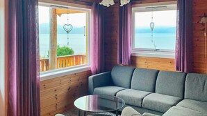 Una habitación acogedora con un sofá gris, una mesita redonda y vistas al mar a través de amplias ventanas con cortinas moradas.