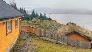Una cabaña de madera con un camino de piedra que lleva a una terraza también de madera, con vistas a un lago y montañas al fondo.