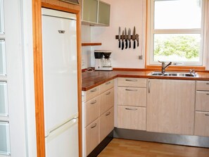 A kitchen with wooden cabinets, a refrigerator, a coffee maker, and a window with a view of greenery. - 6 Person Holiday Home in Lokken (Lokken)