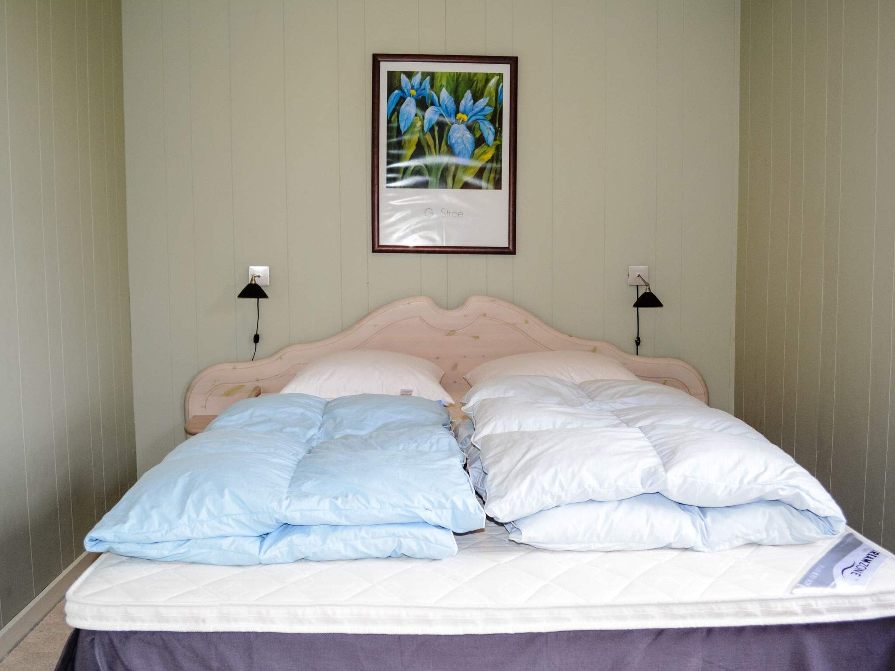 Ein Bett mit weißer Matratze, einem pinken Kopfteil und blau-weißen Kissen. Darüber an der Wand hängt ein gerahmtes Bild von Blumen.