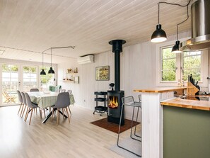 A modern kitchen with a wood-burning stove, a dining area with a table and chairs, and a view of the outdoors through large windows.