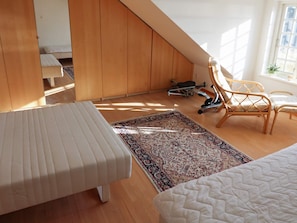 Un dormitorio con techo inclinado, una cama, un banco, una silla, una bicicleta estática pequeña y una alfombra estampada.