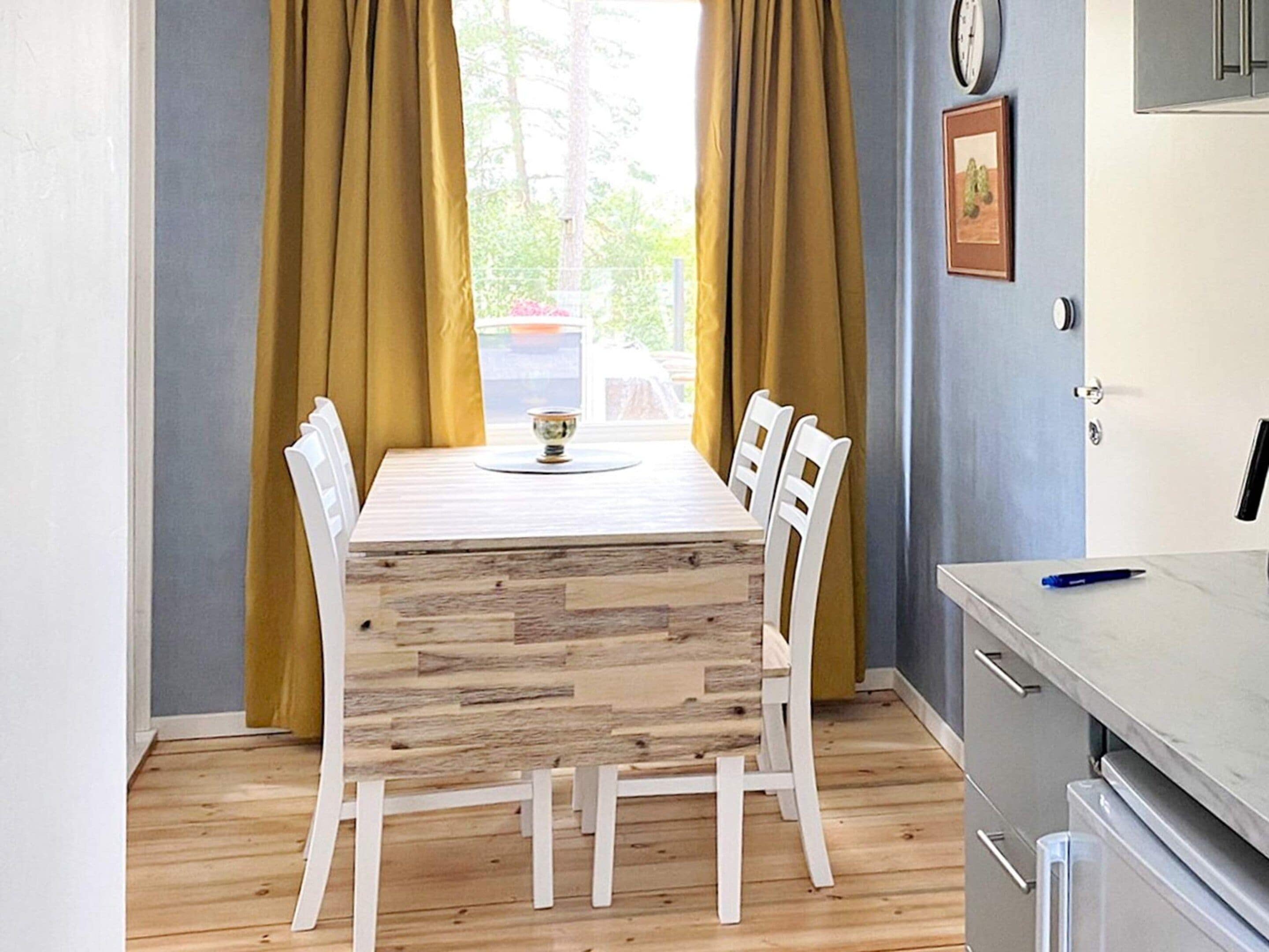 A dining area with a wooden table and white chairs, a window with curtains, a clock, and a kitchen area with cabinets and a countertop.