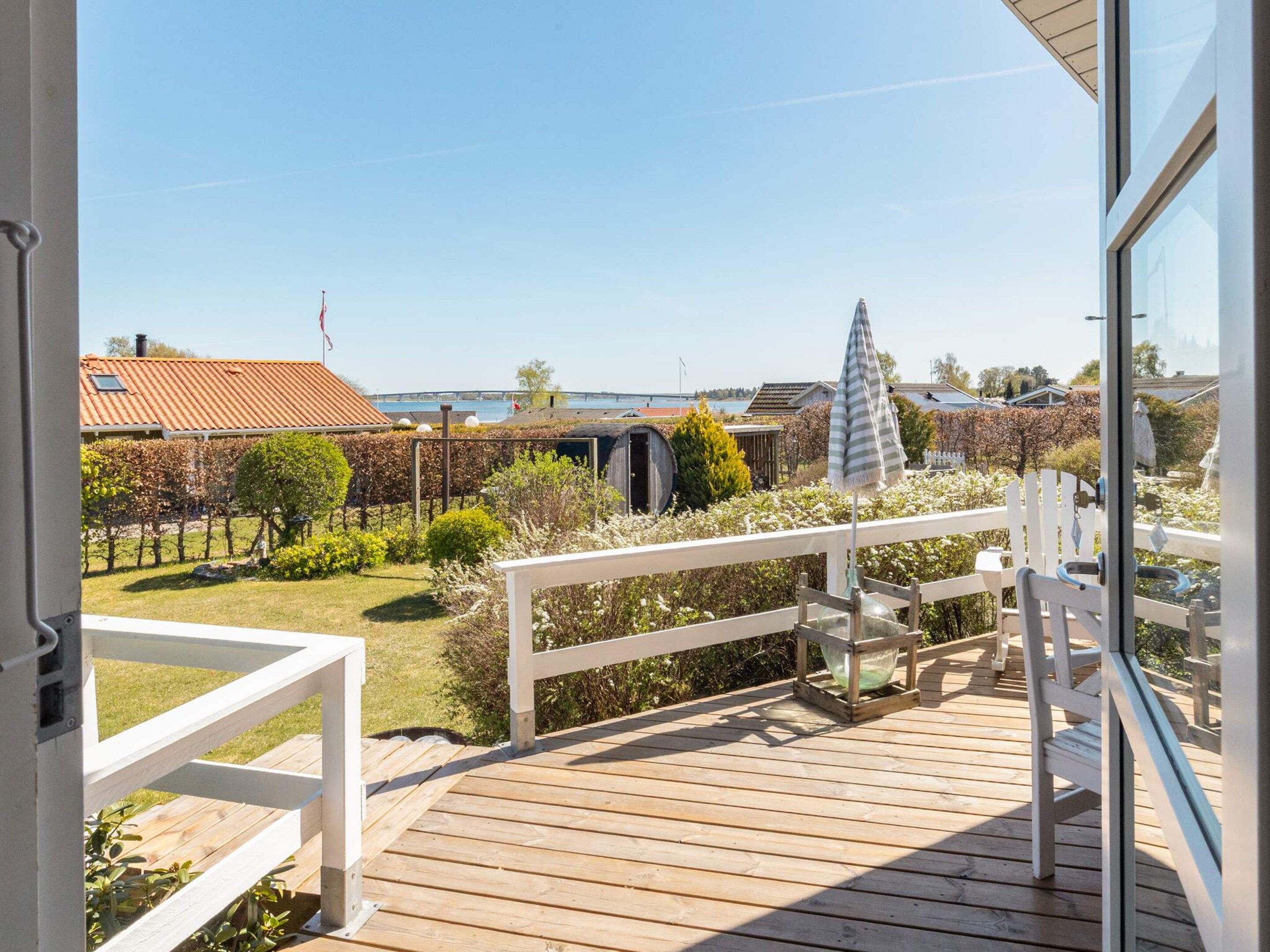A wooden deck with a white railing, a glass door, and a view of a garden with various plants and a distant bridge.