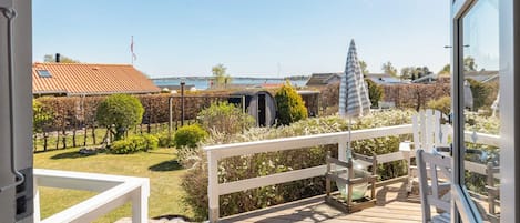 A wooden deck with a white railing, a glass door, and a view of a garden with various plants and a distant bridge.