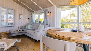 A cozy room with a wooden ceiling, a round wooden table, a sofa, and a window with a view of trees. - Holiday Home in Jægerspris (Jægerspris)