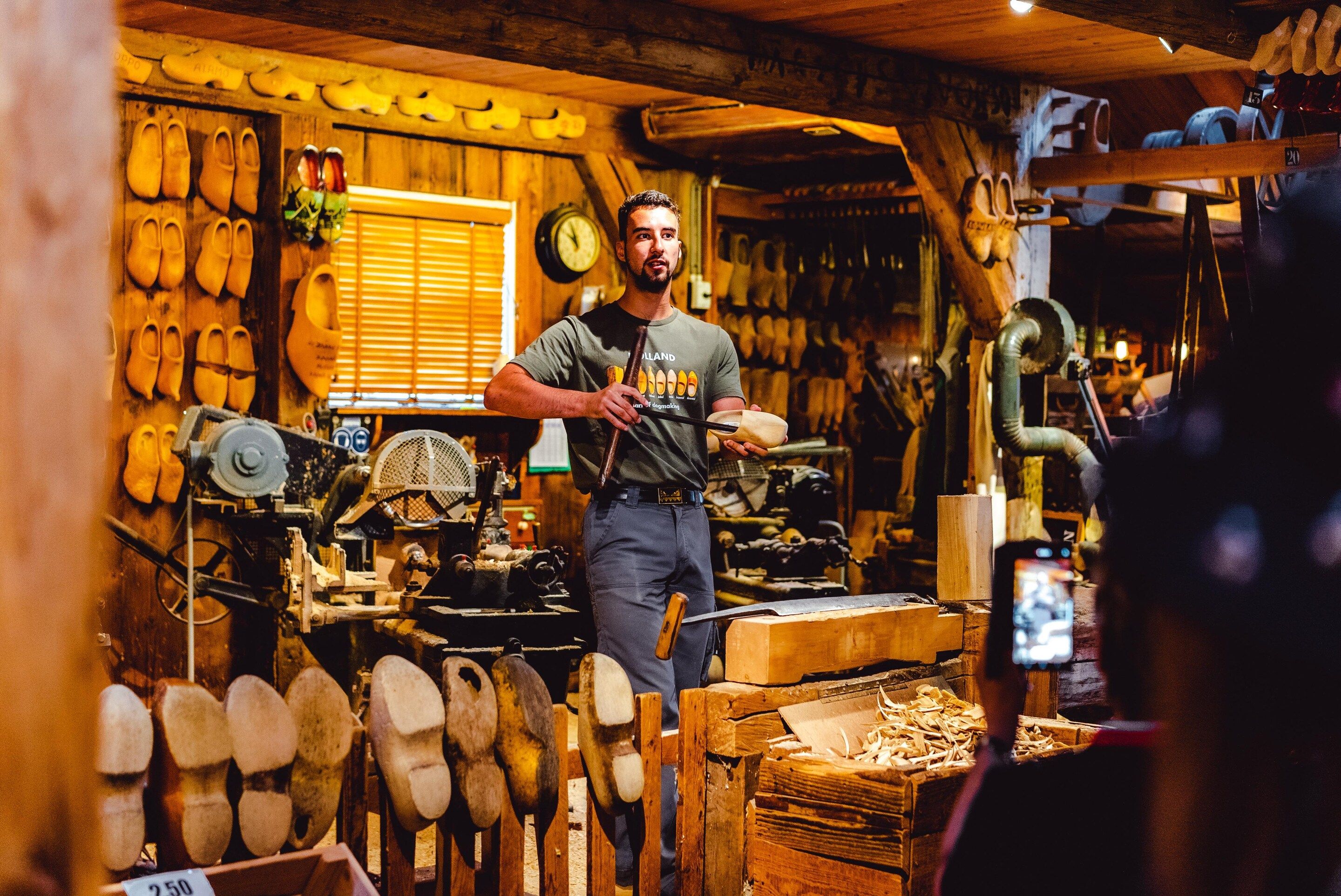 A person in a workshop with woodworking tools and materials, surrounded by wooden shoes and a clock on the wall.