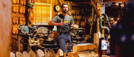 A person in a workshop with woodworking tools and materials, surrounded by wooden shoes and a clock on the wall.