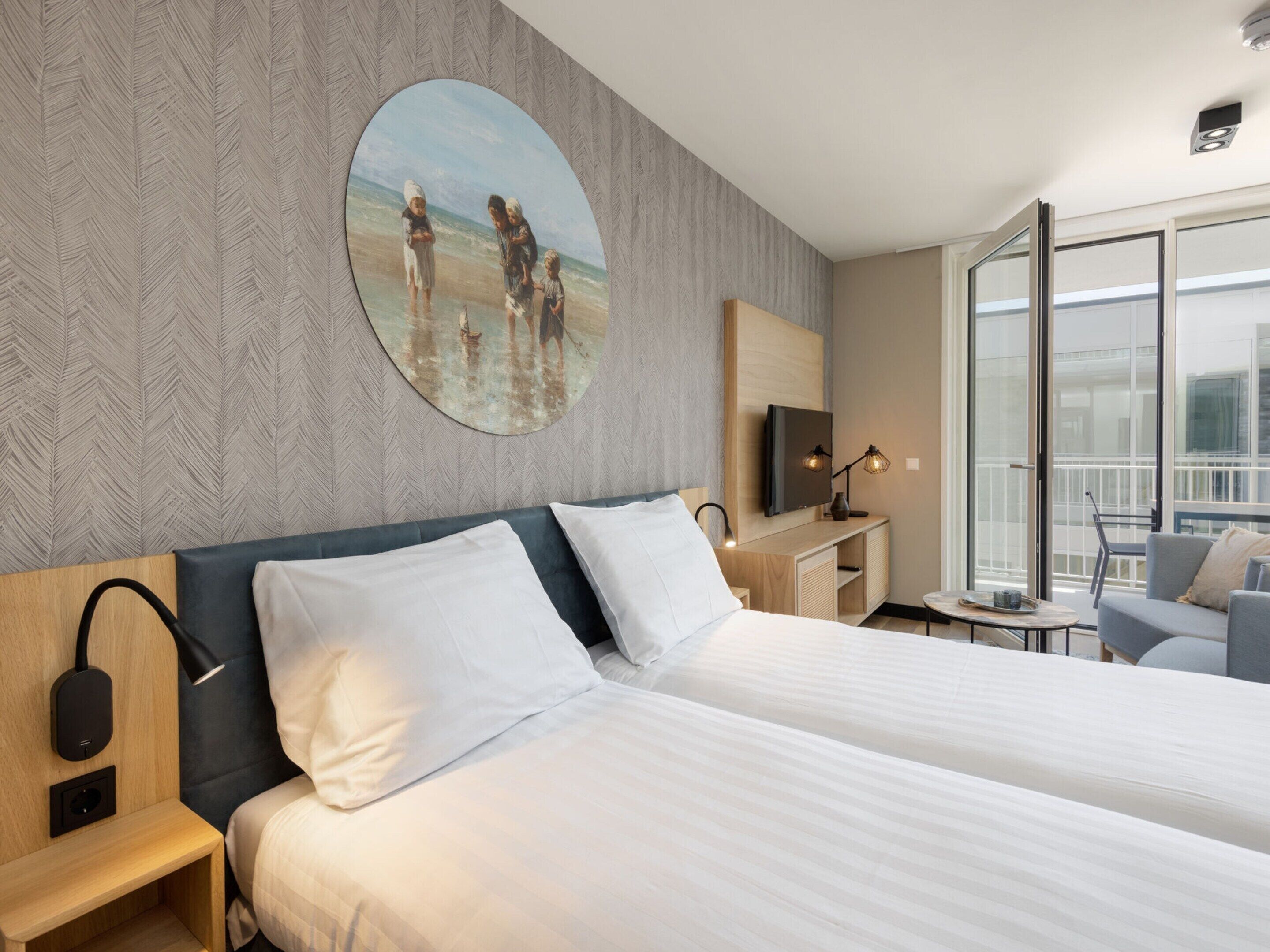 A hotel room with two beds, a large window, a painting on the wall, and a small table with a lamp.