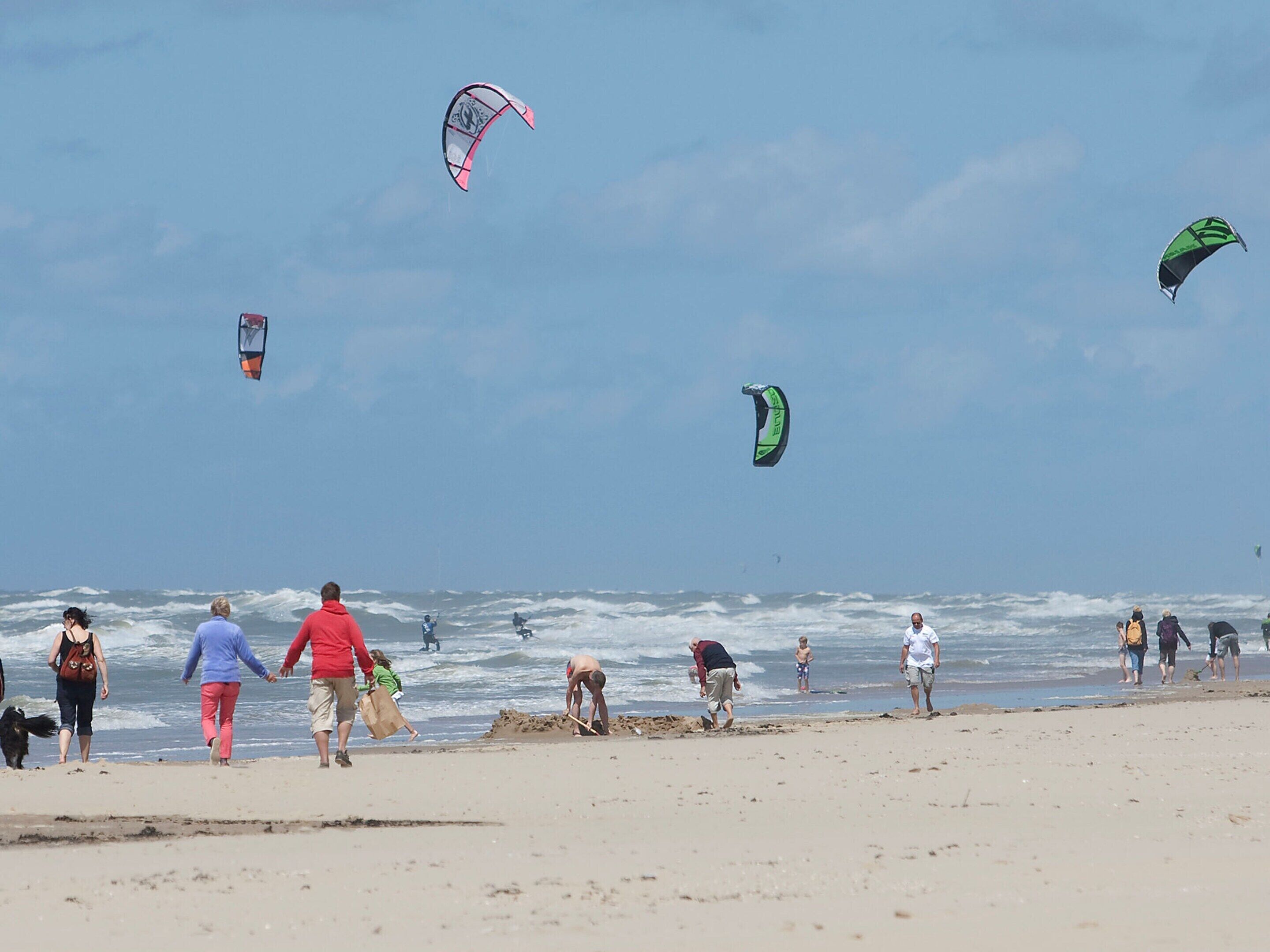 A beach with people walking and playing, and kites flying in the sky.