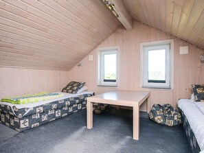 A small, cozy room with wooden walls and a sloped ceiling. There are two windows providing natural light, a bed with patterned bedding, a small table, and a bean bag chair.