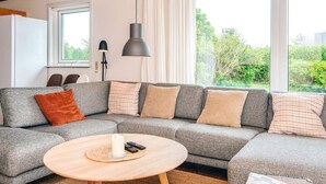 A modern living room with a grey sectional sofa, a round wooden coffee table, and large windows offering a view of greenery outside. - Holiday Home in Odder (Odder)