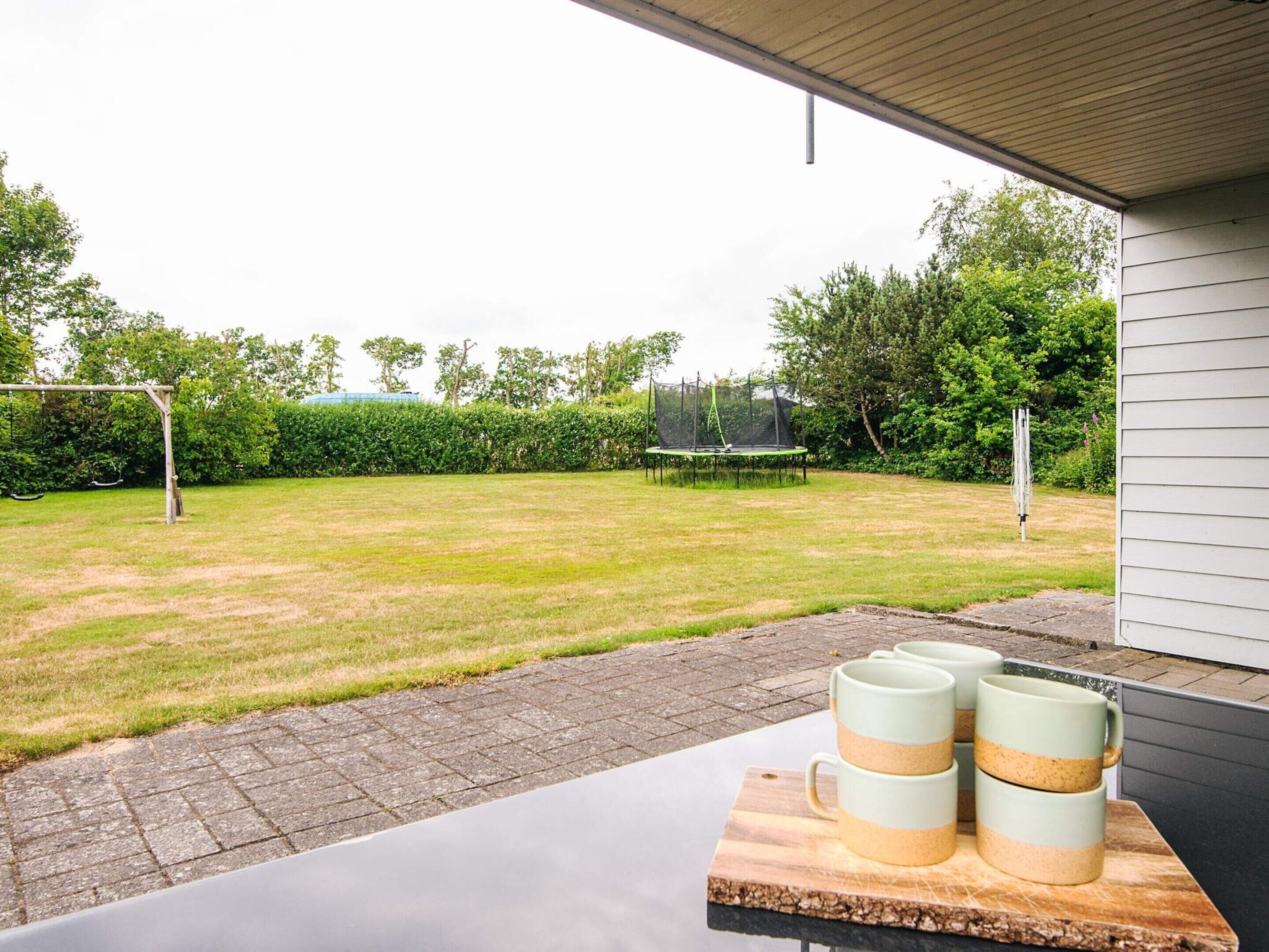 Een terras met een tafel waarop drie mokken staan, een trampoline en een schommelset in een achtertuin.