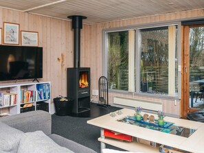 A modern living room with a wood-burning stove, a flat-screen TV, bookshelves, and a large window with a view of trees. - 6 Person Holiday Home in Toftlund (Toftlund)