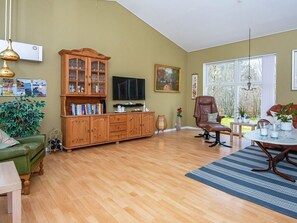 A spacious living room with a wooden cabinet, a television, a green sofa, a dining table with chairs, and a large window providing natural light. - 8 Person Holiday Home in Logstor (Logstor)