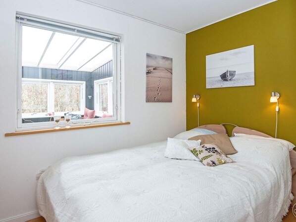 A bedroom with a large window, a bed with white bedding, two wall-mounted lamps, and two framed pictures.